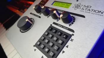 An Elektron SidStation synthesizer with a MIDI controller keyboard in the background